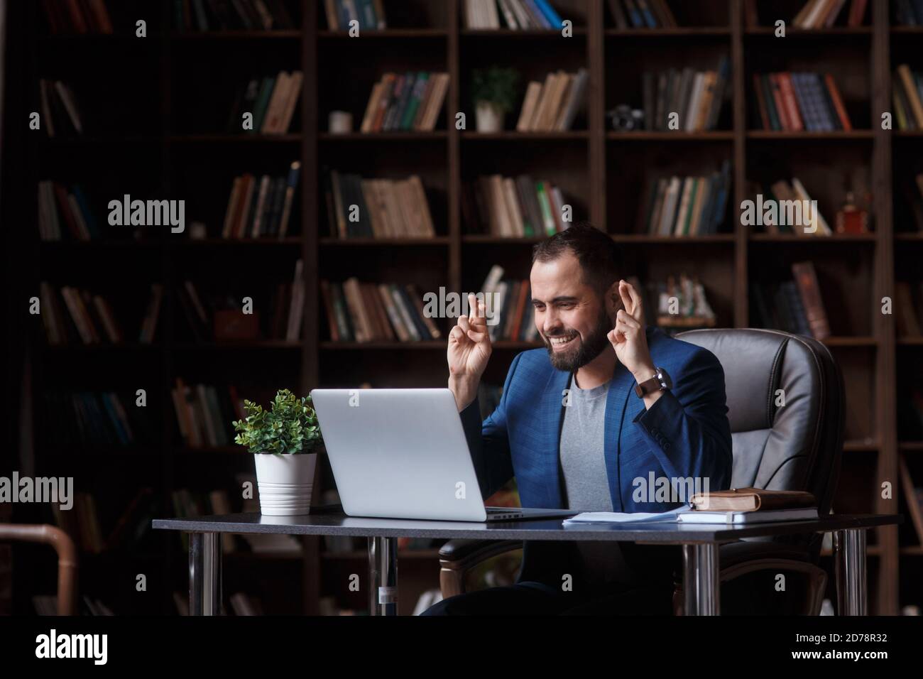 Un giovane uomo d'affari con la bearded nell'ufficio della biblioteca in un vestito blu guarda lo schermo del laptop e le attraversa le dita. Concetto di attesa per Foto Stock