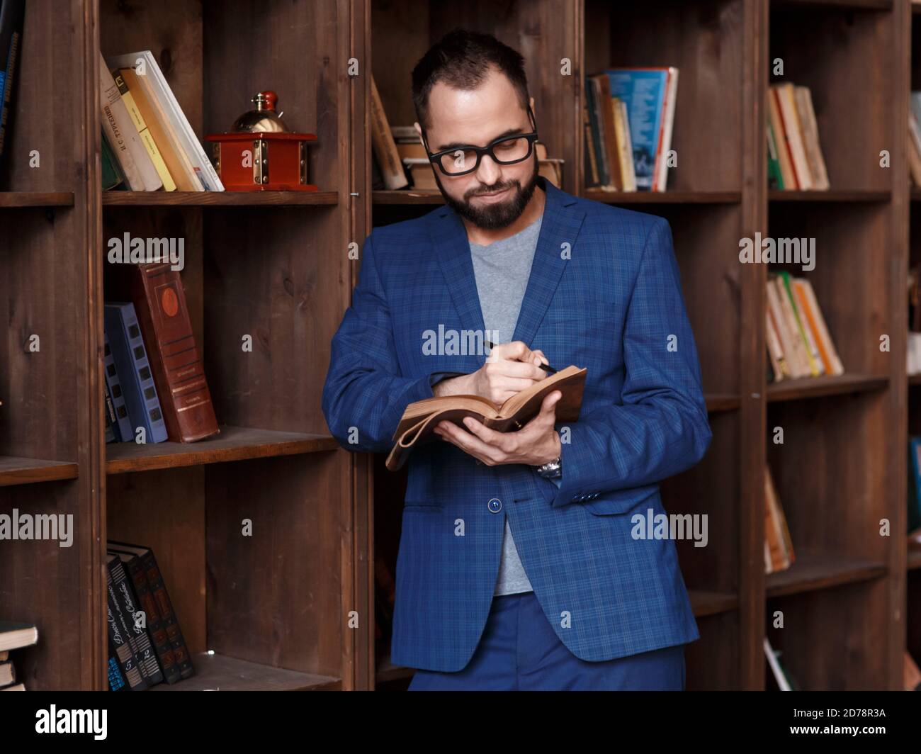 uomo d'affari bearded con gli occhiali in biblioteca con un taccuino in mano sorride. Giovane scrittore Foto Stock