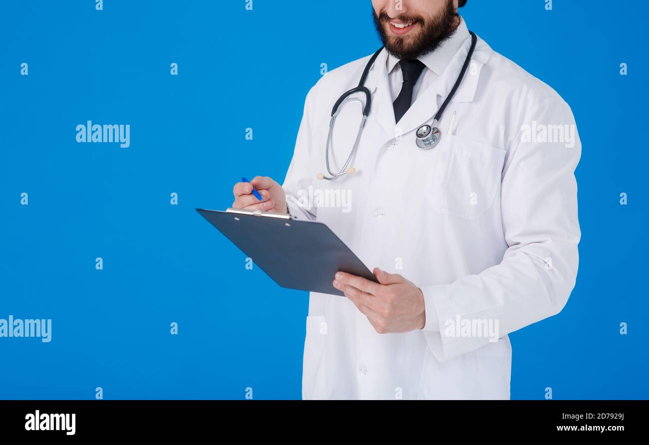 un giovane medico con la bearded in un cappotto medico bianco sopra uno sfondo blu scrive i reclami del paziente su un tablet con documenti e medici Foto Stock