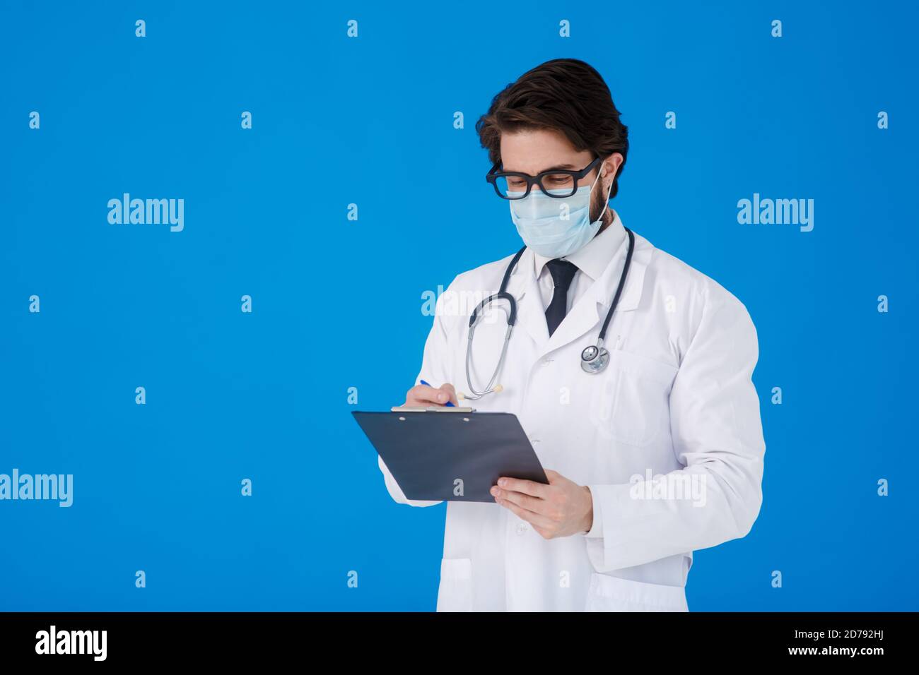 un giovane medico sopportato in un cappotto medico bianco e. una maschera su sfondo blu scrive il paziente reclami su un tablet con documenti e. Foto Stock