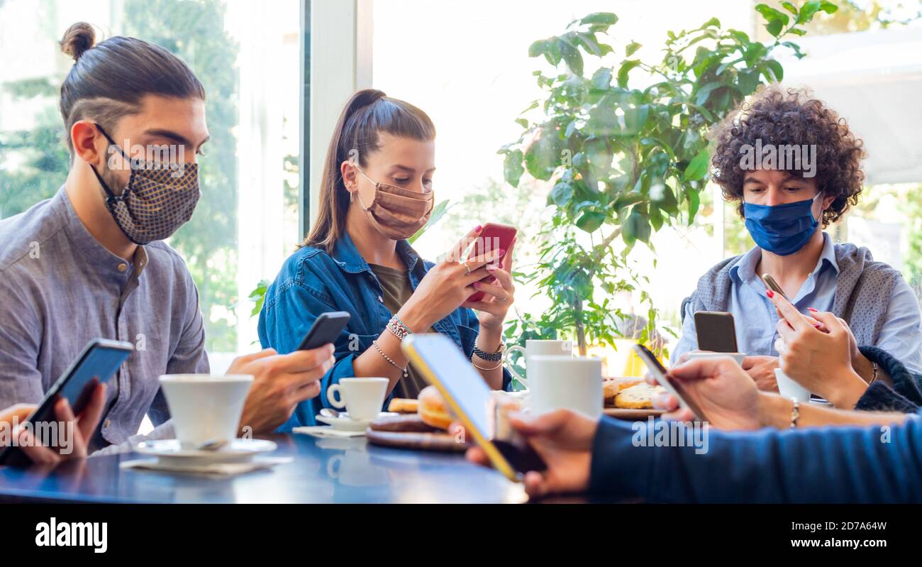 un gruppo di amici sorridenti a colazione in un bar mentre interagiscono con la tecnologia. i compagni bevono caffè e mangiano mentre usano gli smartphone. Foto Stock