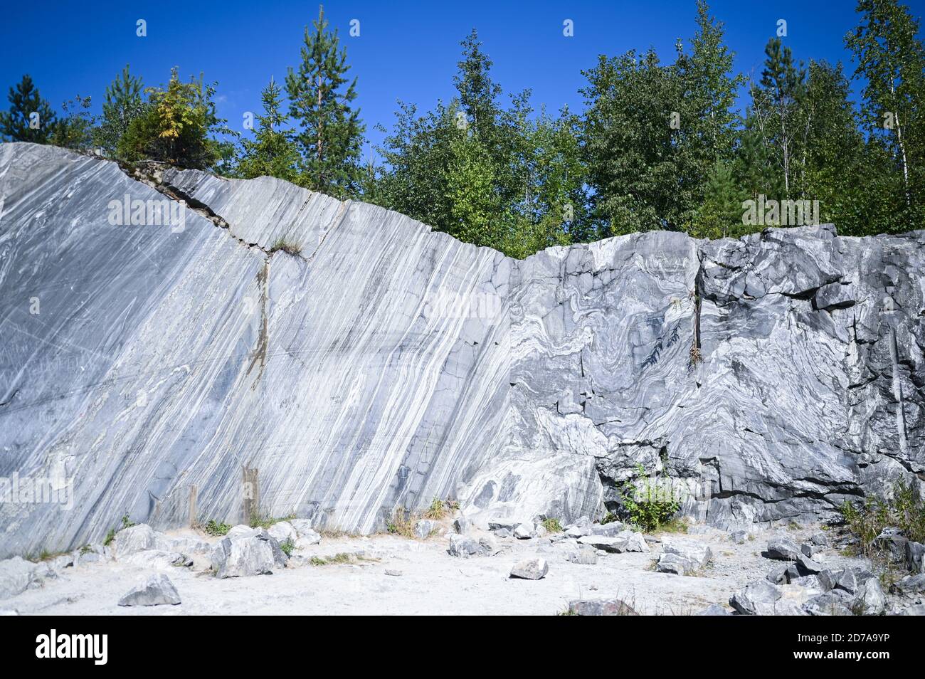 Superficie grezza di marmo bianco e grigio tagliata da roccia in ambiente naturale con alberi e cielo blu. Marmo blu tessitura carriera. Pietra di fondo, materiale industriale per edifici e interni. Foto Stock