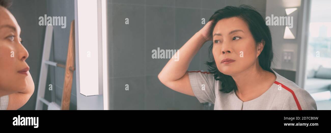 Donna matura asiatica di mezza età che tocca i suoi capelli guardando se stessa in bagno specchio panoramico banner stile di vita casa. Senior chinese signora capelli Foto Stock