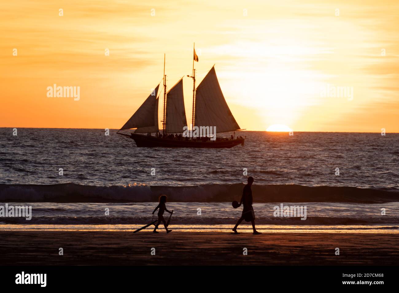 Padre e figlio camminano lungo Cable Beach al tramonto mentre una nave a vela porta i turisti in una crociera al tramonto. Foto Stock