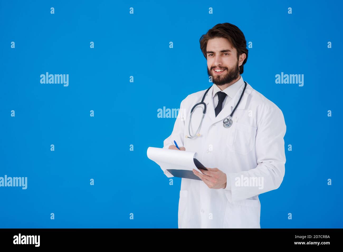 un giovane medico con la bearded in un cappotto medico bianco sopra uno sfondo blu scrive i reclami del paziente su un tablet con documenti e medici Foto Stock
