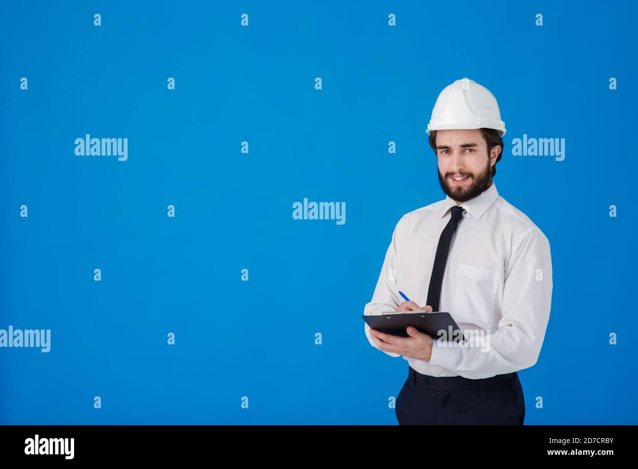 Un giovane uomo portatore di costruzioni in una camicia bianca e casco da costruzione su sfondo blu. Ingegnere industriale-architetto tenere appunti Foto Stock