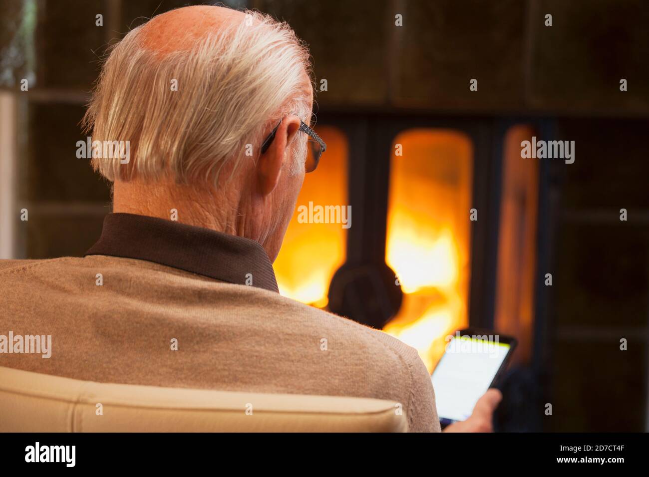 Uomo anziano seduto da un camino e guardando un smartphone: messa a fuoco selettiva sulla testa Foto Stock