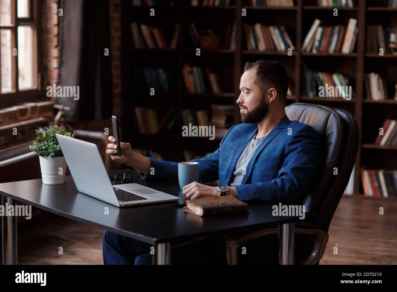 Un giovane uomo d'affari in una tuta blu nell'ufficio della biblioteca sta parlando su un collegamento video sul telefono. Lo spazio di lavoro dell'uomo d'affari - Foto Stock