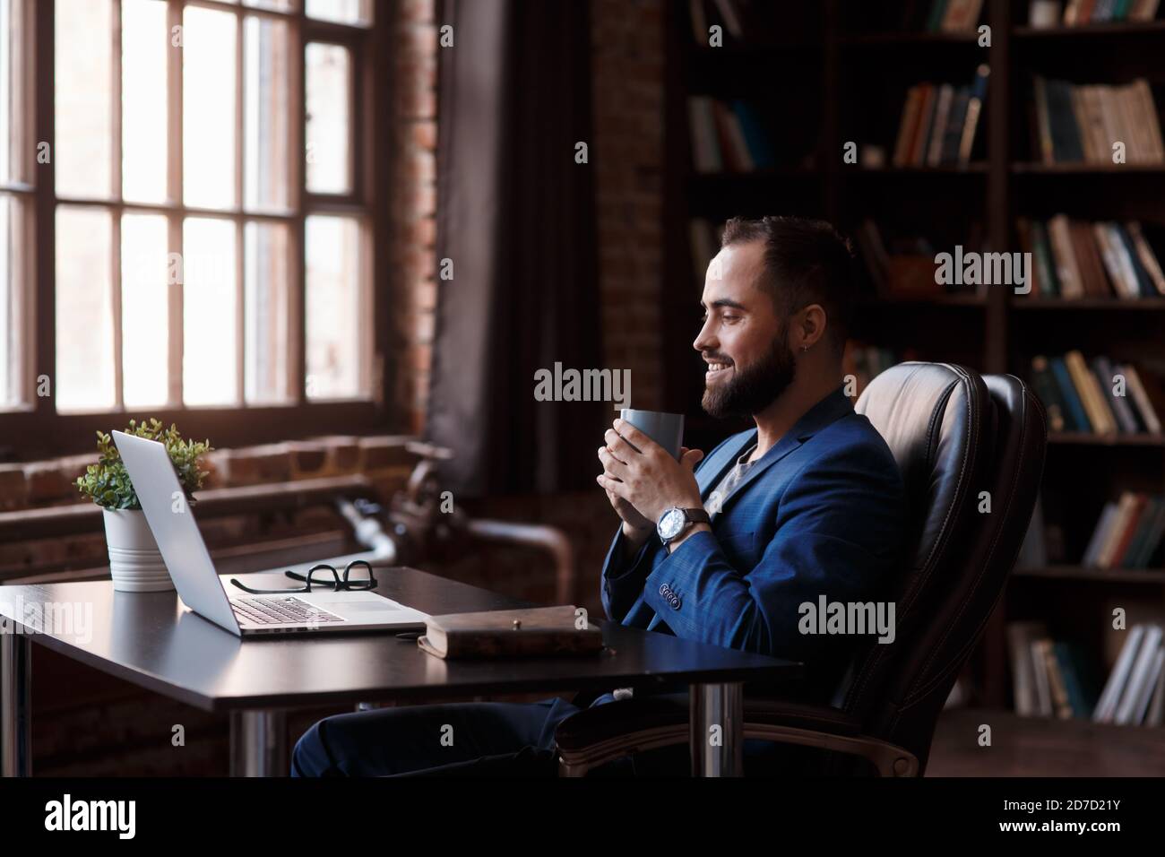 Caffè fresco al lavoro. Giovane sicuro in abbigliamento casual elegante che lavora su un computer portatile e beve una bevanda calda mentre si siede al lavoro Foto Stock