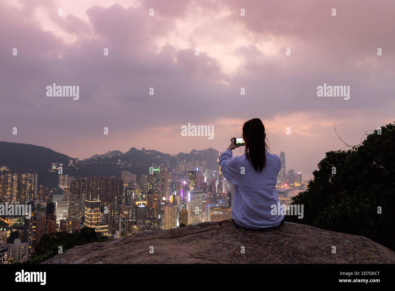 Silhouette di ragazza che siede al rock e scatta foto della vista notturna della città Skyline di Hong Kong Island (a sud di Victoria Harbour) dalla collina di Braemar, Foto Stock