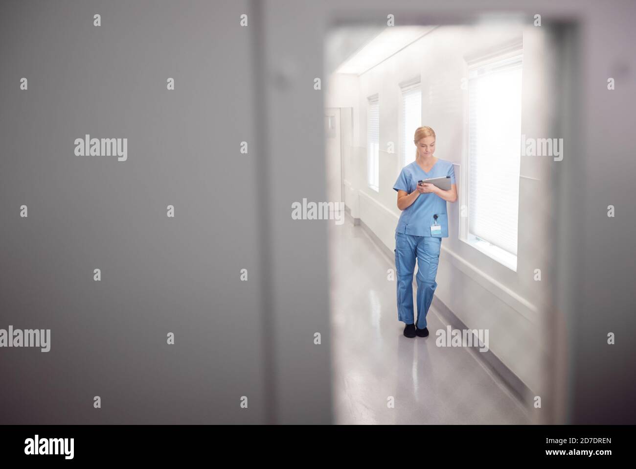 Dottore femminile che indossa scrub nel corridoio dell'ospedale che utilizza la tavoletta digitale Vista attraverso la finestra nella porta Foto Stock