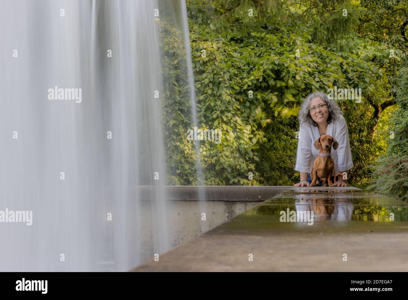 Sorridente donna messicana con il suo dachshund seduto tranquillamente guardando la macchina fotografica, accanto ad una cascata da una grande fontana con lussureggiante vegetazione verde i Foto Stock