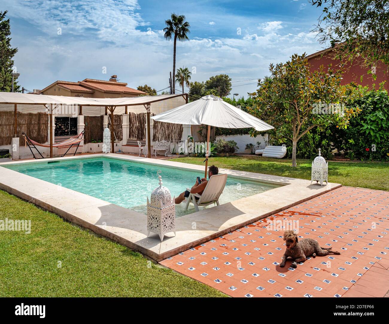 Uomo non identificato che si rilassa in una villa di vacanza con il suo  cane in una piscina con sdraio abbronzante, piscina con sedie, ombrellone,  erba, giardino, portico e gazebo Foto stock -