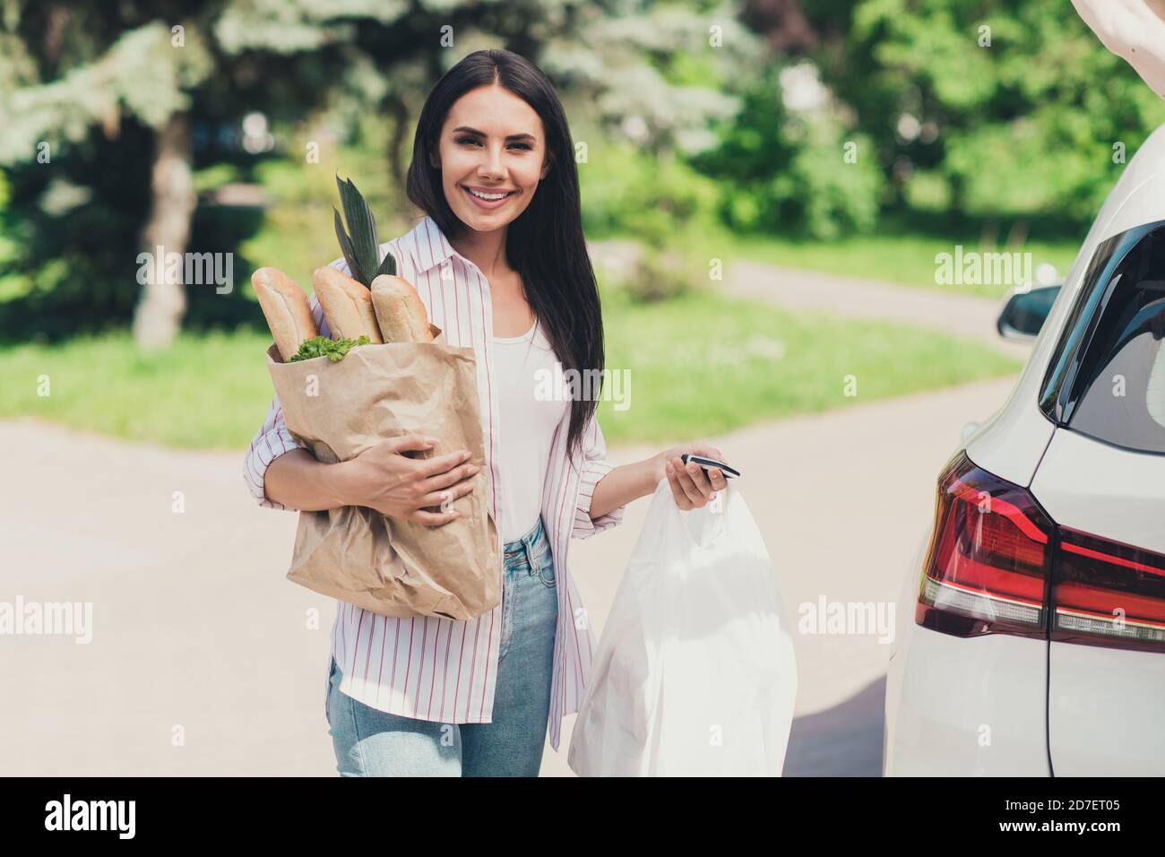 Foto di positivo affascinante ragazza andare shopping mercato commerciale acquistare cibo verdure pane porro per la famiglia cena a piedi auto tenere le chiavi della borsa aprono il vano bagagli in città Foto Stock