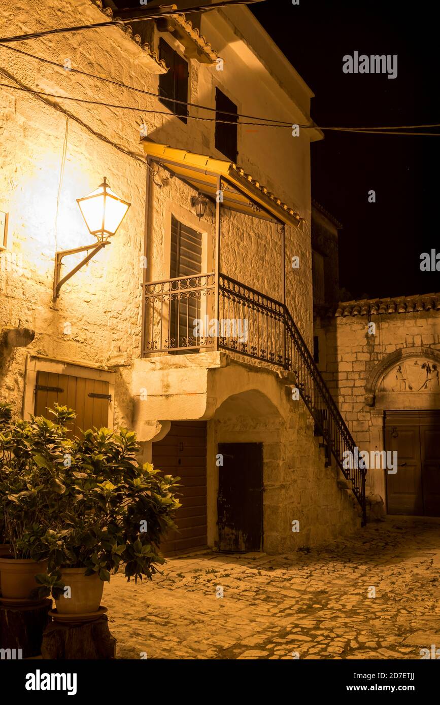 Strada deserta con casa in pietra con scale e marciapiede lastricato in zona medievale storica della città di Trogir, Croazia illuminata da lanterne di notte Foto Stock