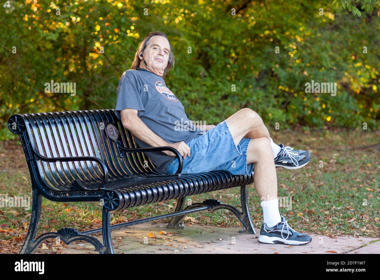Frederick, MD, USA 10/14/2020: L'uomo di mezza età dai capelli lunghi si rilassa su una panchina del parco dopo un'escursione. Ruota la testa mentre è seduto a gambe incrociate Foto Stock