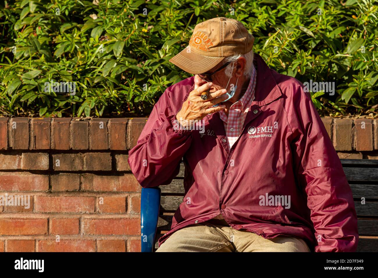 Frederick, MD, USA 10/14/2020: Un uomo caucasico anziano è seduto da solo su una panchina di strada e fuma un sigaro spesso. Mette la sua maschera contro CO Foto Stock