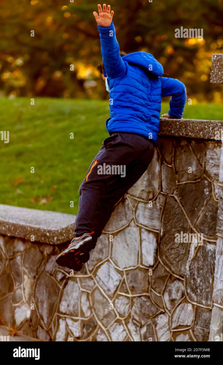 Un bambino iperattivo sta saltando da un muro alto mentre sta correndo via. Il ragazzo coraggioso indossa pantaloni da atletica, sneakers e un cappotto con cappuccio. È pieno di it Foto Stock
