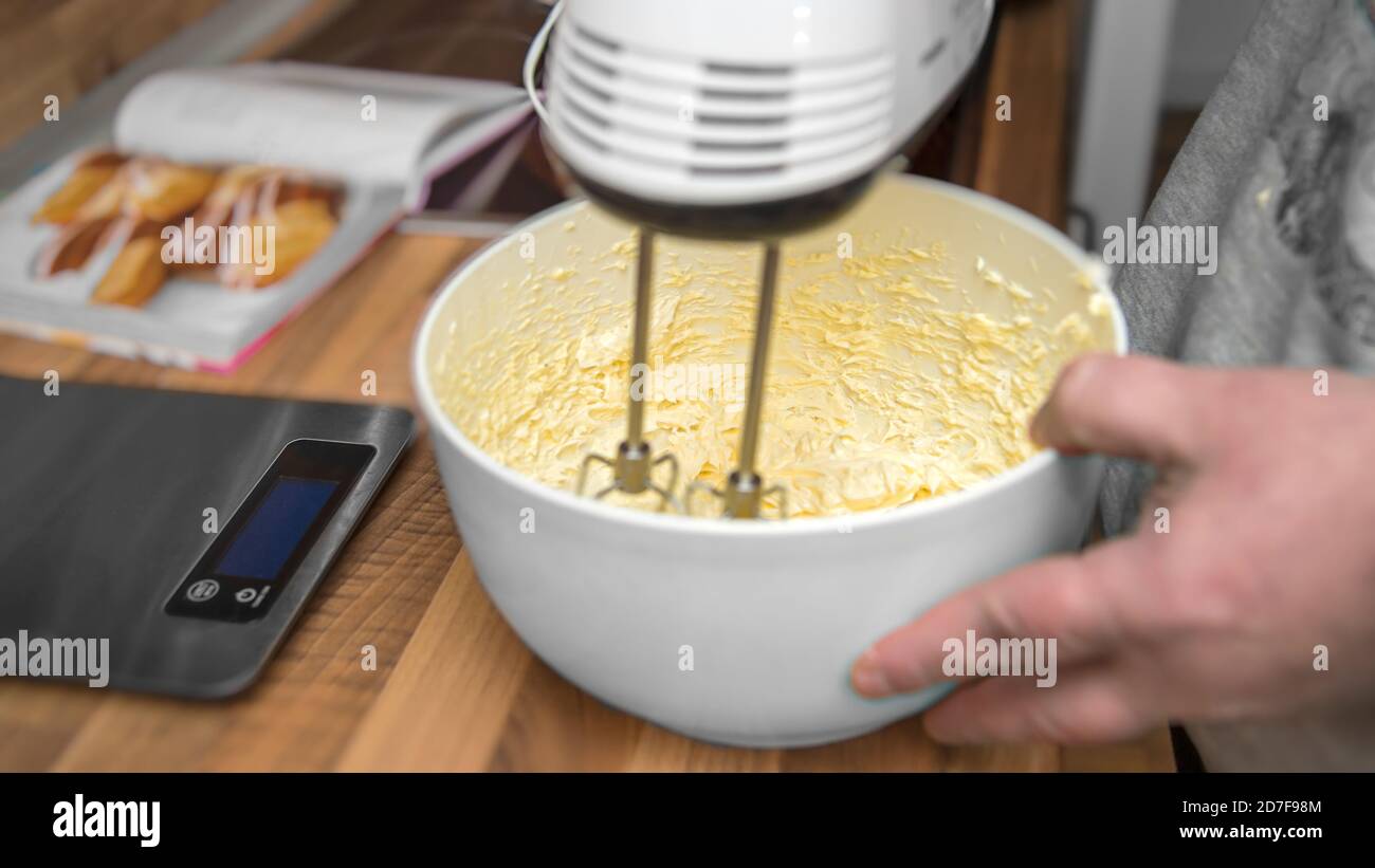 Un uomo tiene un miscelatore elettrico della mano. Mescola burro, uova e zucchero. Viene preparata una pasta frolla. Primo piano dalla cucina. Foto Stock