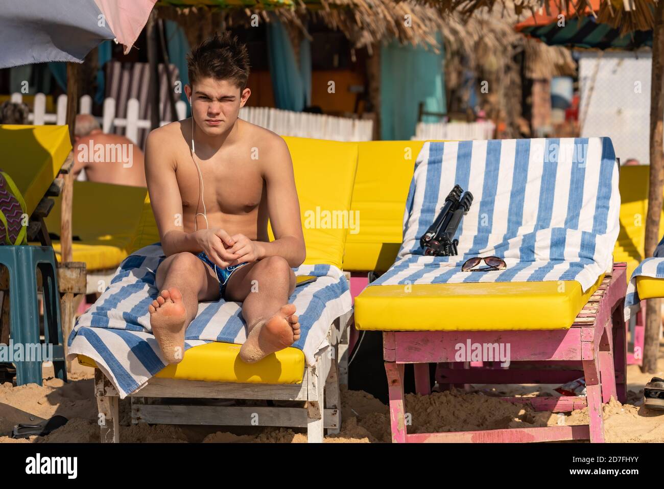 Carino bel ragazzo caucasico che ascolta musica con un auricolare Seduto sul lettino del sole sulla spiaggia tropicale con il volto scettico espressione - solo arri Foto Stock