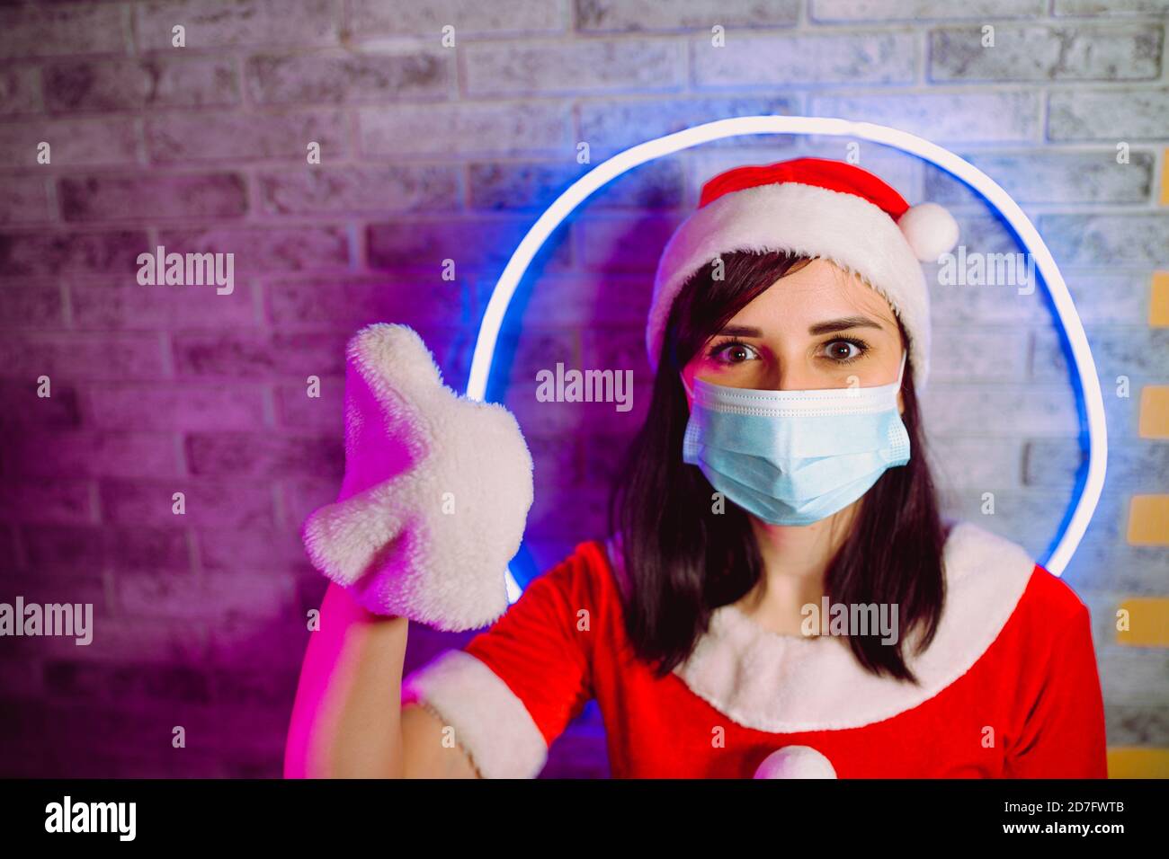 Donna in maschera medica, tuta Babbo Natale mostra gesto di approvazione contro parete illuminata. Donna in cappello di Natale e maschera protettiva. Concetto di Foto Stock