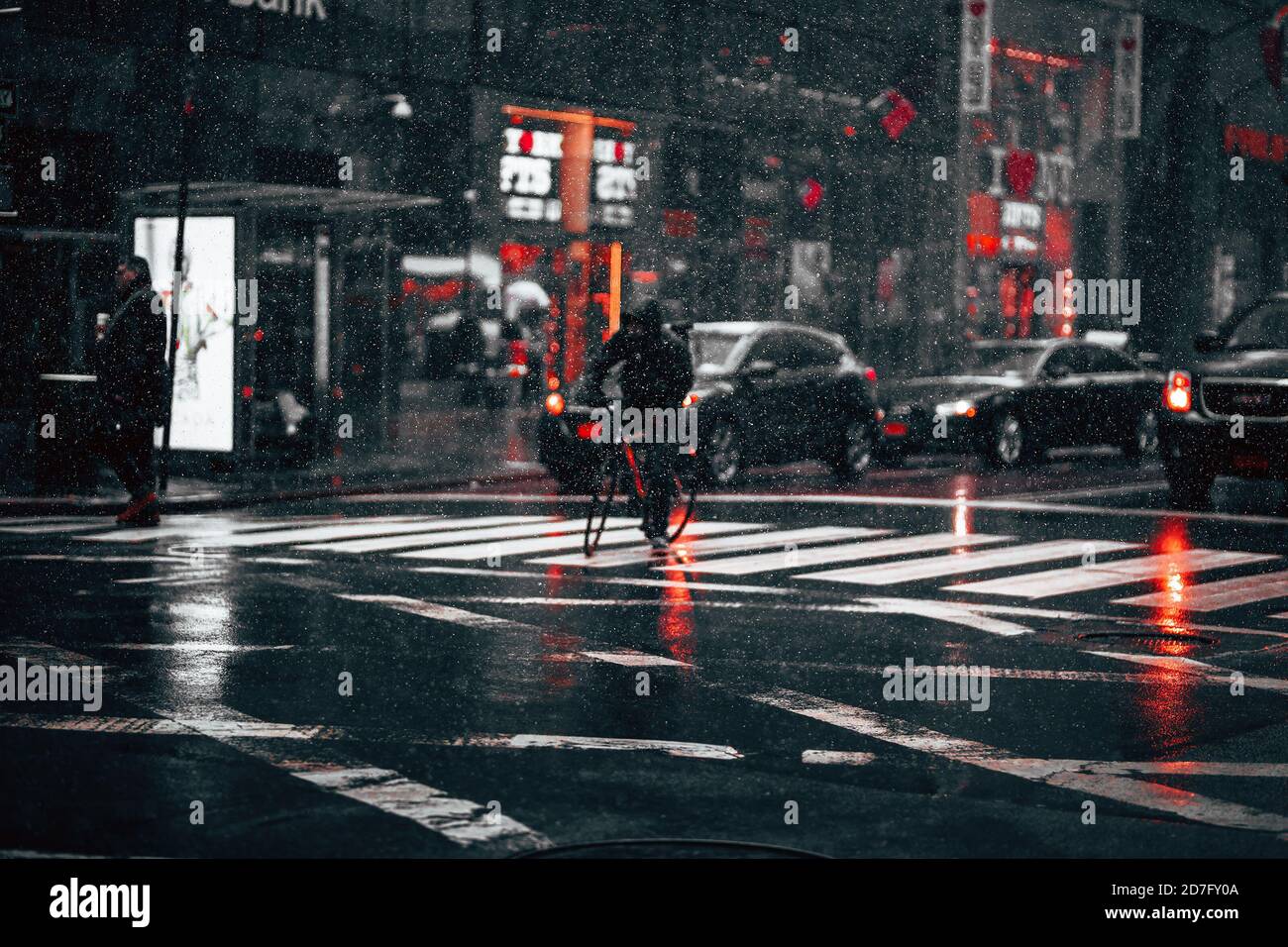 Piovendo strada con semafori di notte. Vita cittadina in un giorno piovoso. Gocce di pioggia si fermavano in movimento contro luci di auto, biciclette e persone che camminavano Foto Stock