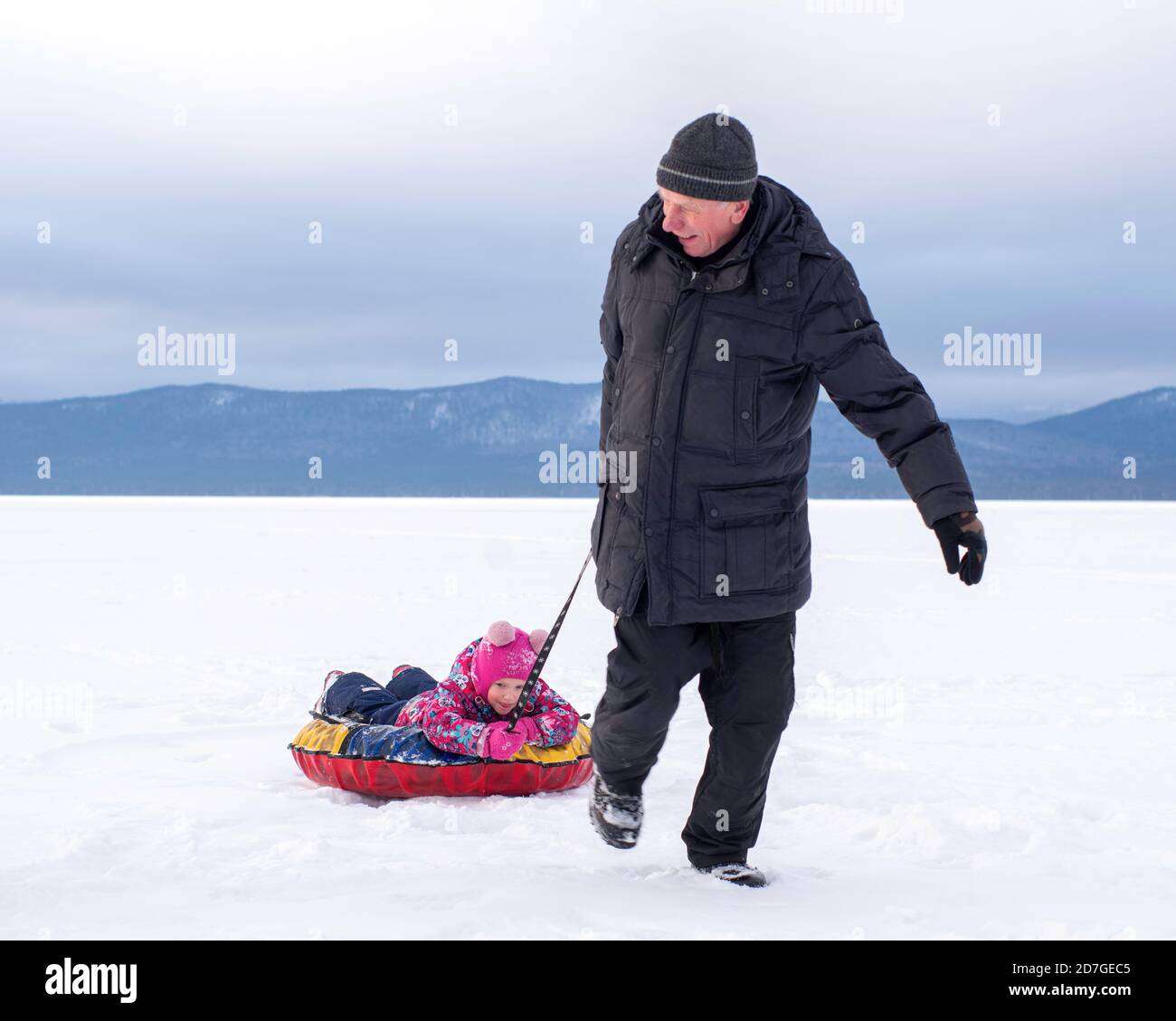 Un uomo, un anziano, sta portando una ragazza felice su una slitta gonfiabile, che passa attraverso la neve bianca. Concetto di giochi invernali all'aperto Foto Stock