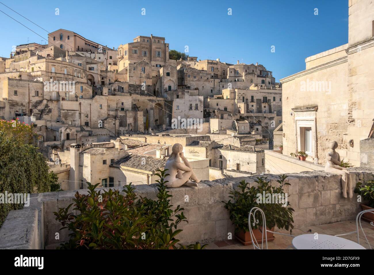 Un piccolo cortile patio con tavoli e due figure scultoree d'arte moderna con il borgo alle spalle nell'antica città sassi di Matera, Italia. Foto Stock