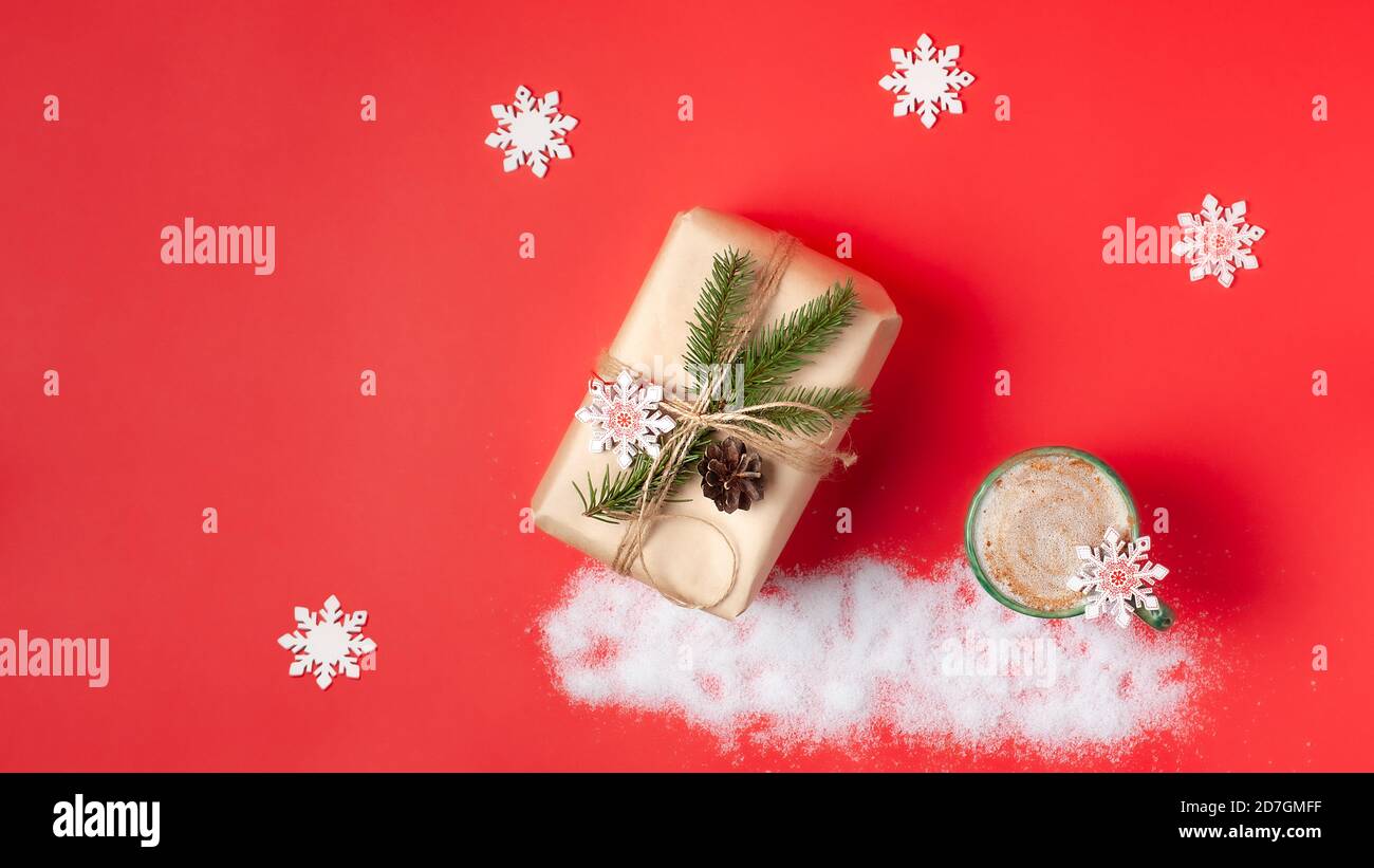 Bella scatola regalo avvolta in carta artigianale e decorata con ramo cipresso su sfondo di carta rossa. Tazza con cappuccino nelle vicinanze. Disposizione piatta, spazio di copia Foto Stock