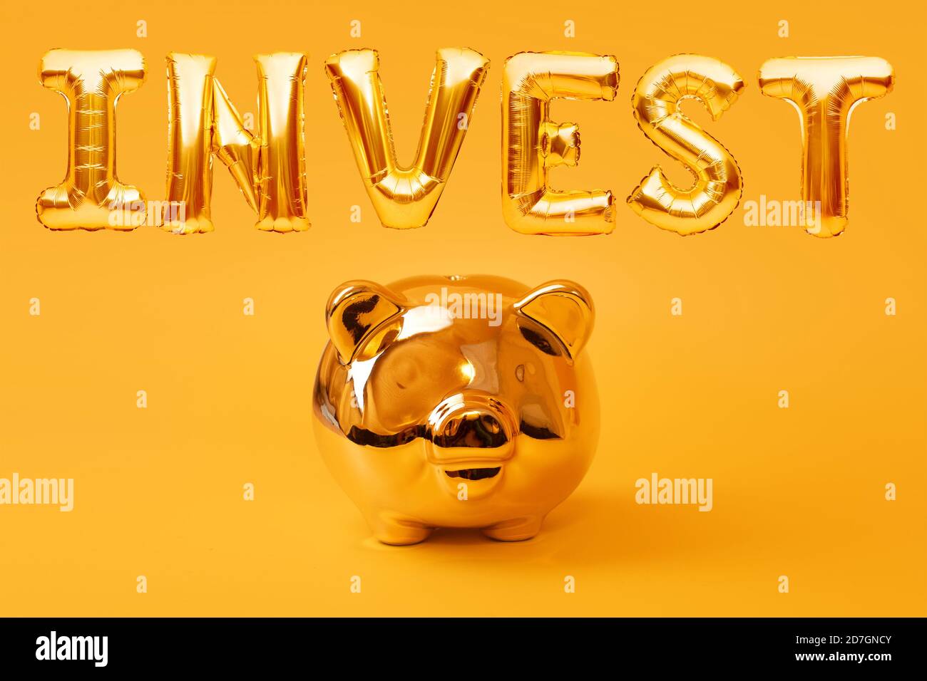 Golden piggy banca su sfondo giallo con la parola d'oro INVESTIRE fatto di palloncini gonfiabili. Investimento e concetto bancario. Risparmio di denaro Foto Stock