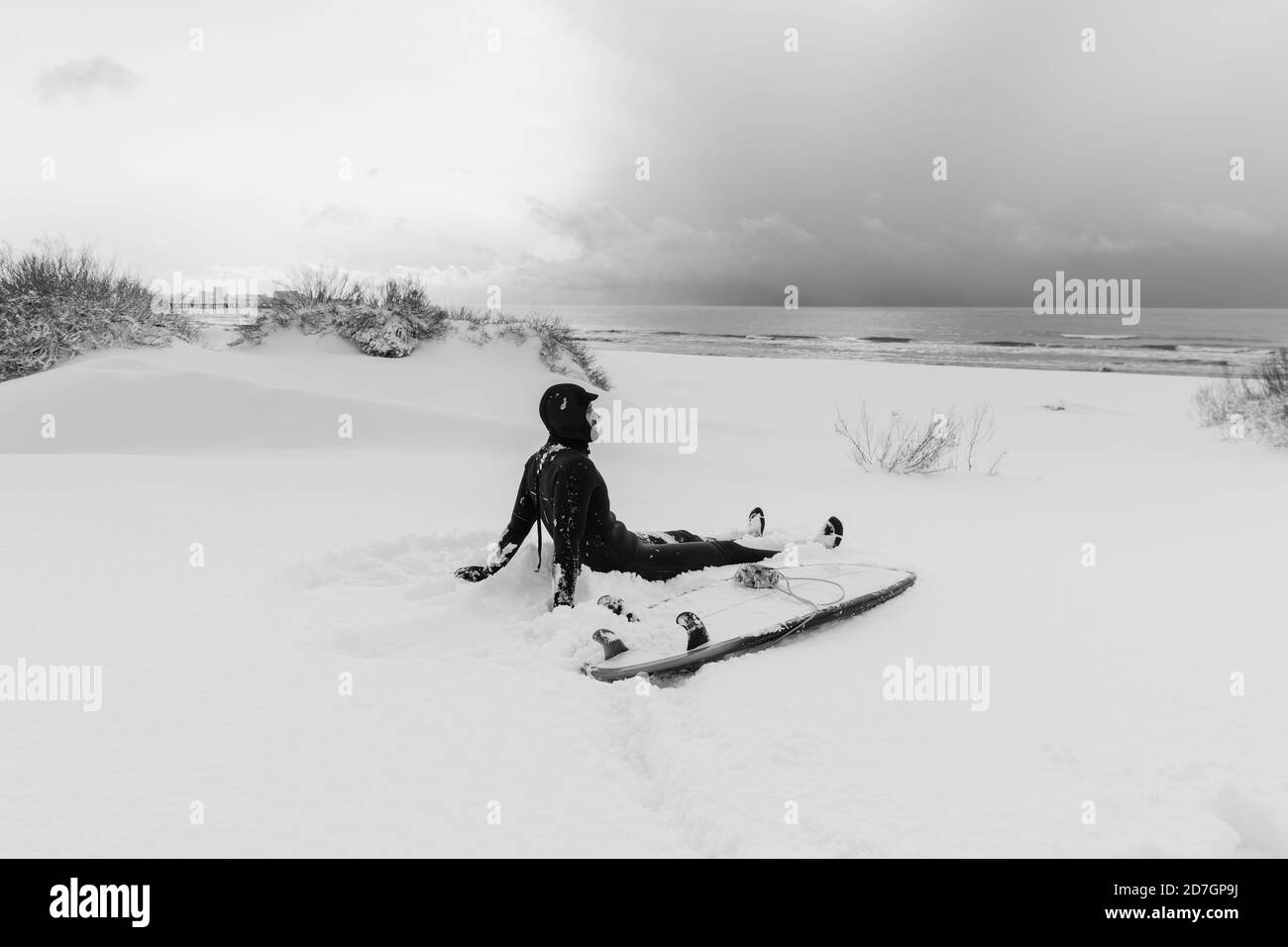 Inverno freddo e surfista sedersi sulla spiaggia innevata con surf e vedere a onde. Giornata invernale con surfista in muta. Foto Stock