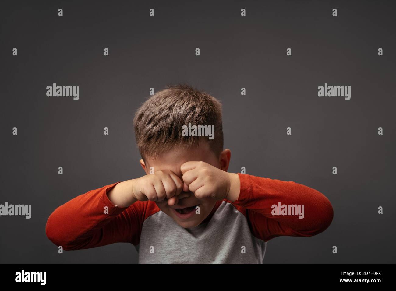 Il ragazzino piangente esprime tristi emozioni. Il bambino sconvolto sfrega gli occhi con i pugni isolati su sfondo grigio. Concetto di problema Foto Stock