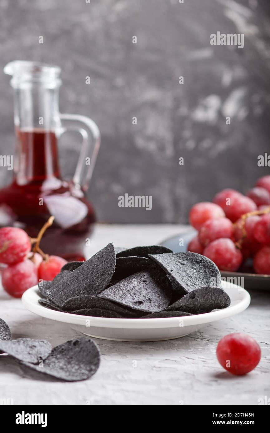 Patatine nere con carbone, aceto balsamico in vetro, uva rossa su un piatto di ceramica blu su fondo grigio. Vista laterale, primo piano, se Foto Stock