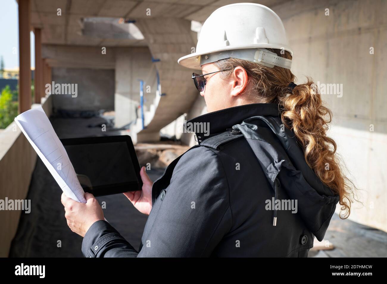Vista posteriore di una giovane donna architetto che indossa un casco di sicurezza, che tiene piani di pavimento in carta e verifica di un dispositivo digitale in un cantiere Foto Stock