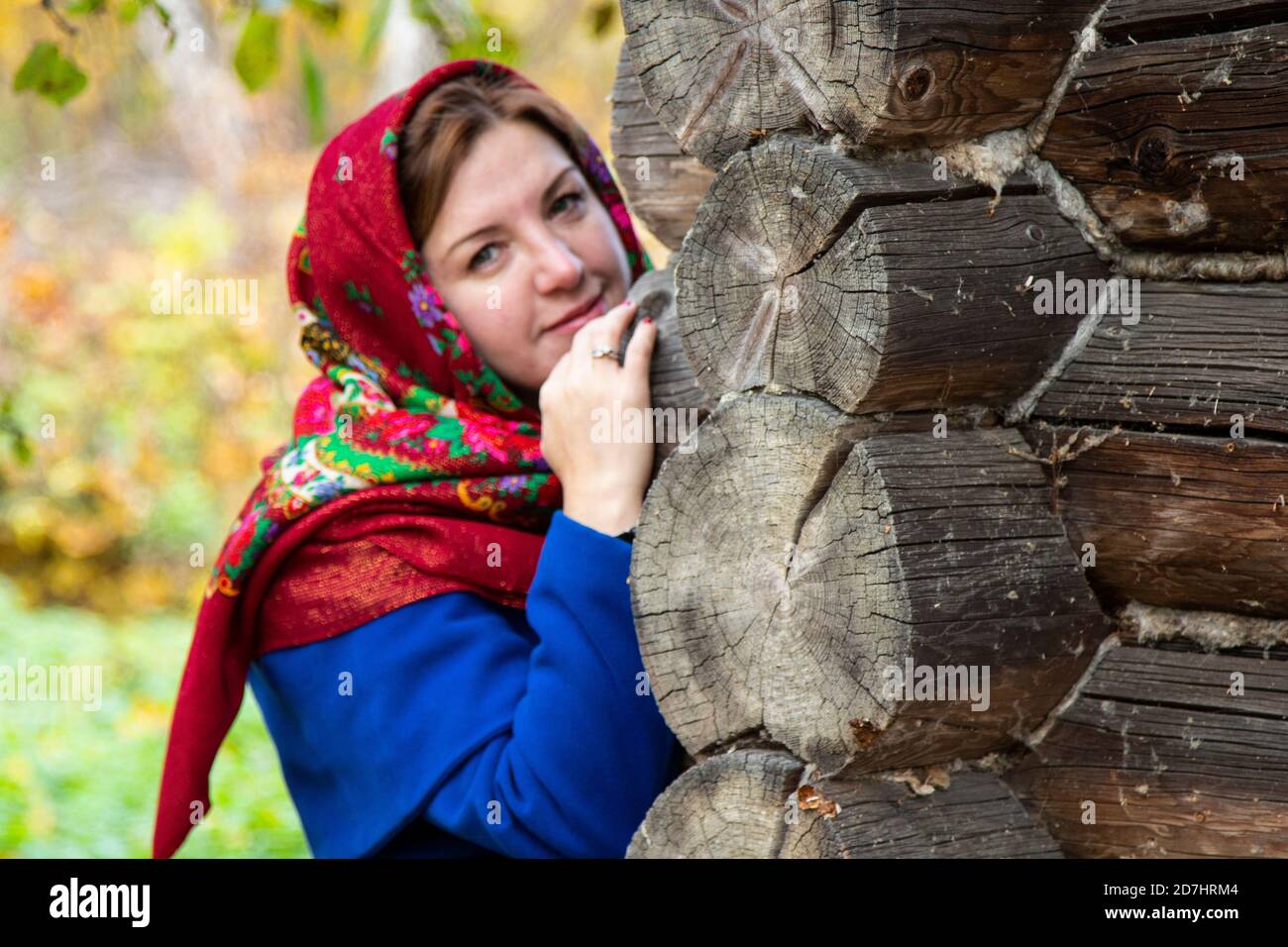 Una giovane donna in un tradizionale copricapo russo si affaccia dietro  l'angolo di una casa di legno. Fazzoletto Pavlovsk Foto stock - Alamy