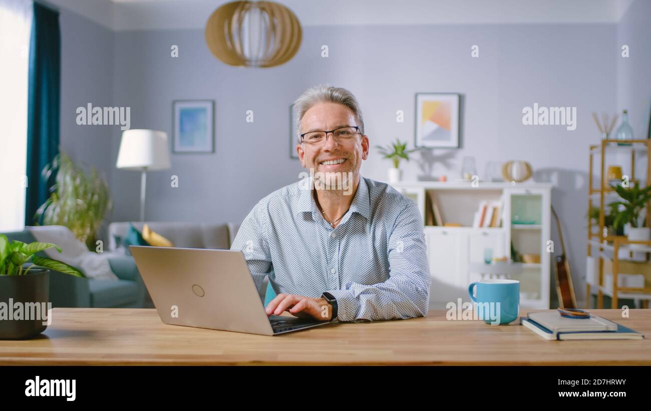 Bell'uomo di mezza età usa il computer portatile e sorride sulla fotocamera. È nel salotto accogliente ed elegante. Felice uomo di successo funziona sul computer Foto Stock