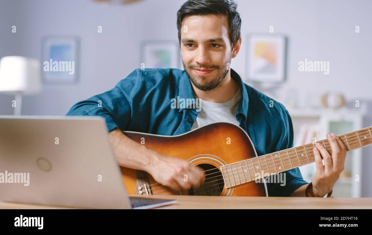 Il giovane uomo di talento gioca la chitarra mentre si siede alla sua scrivania, leggendo i Chords da un laptop, facendo il video blogging in linea. Uomo che pratica la musica della chitarra Foto Stock