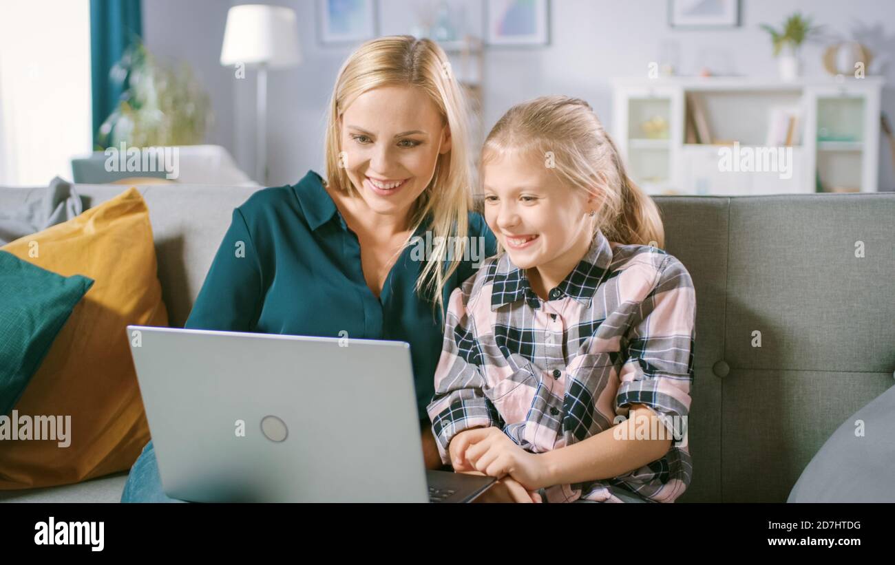 Bella giovane mamma e il suo carino piccolo daugther utilizzare il laptop mentre si siede su un divano a casa. La famiglia trascorre il tempo insieme guardando i video e. Foto Stock