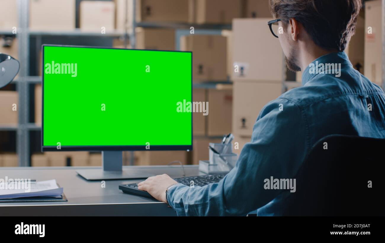 Il responsabile dell'inventario del magazzino lavora su un computer con schermo verde mentre è seduto alla sua scrivania. Negli scaffali di sfondo pieni di scatola di cartone Foto Stock
