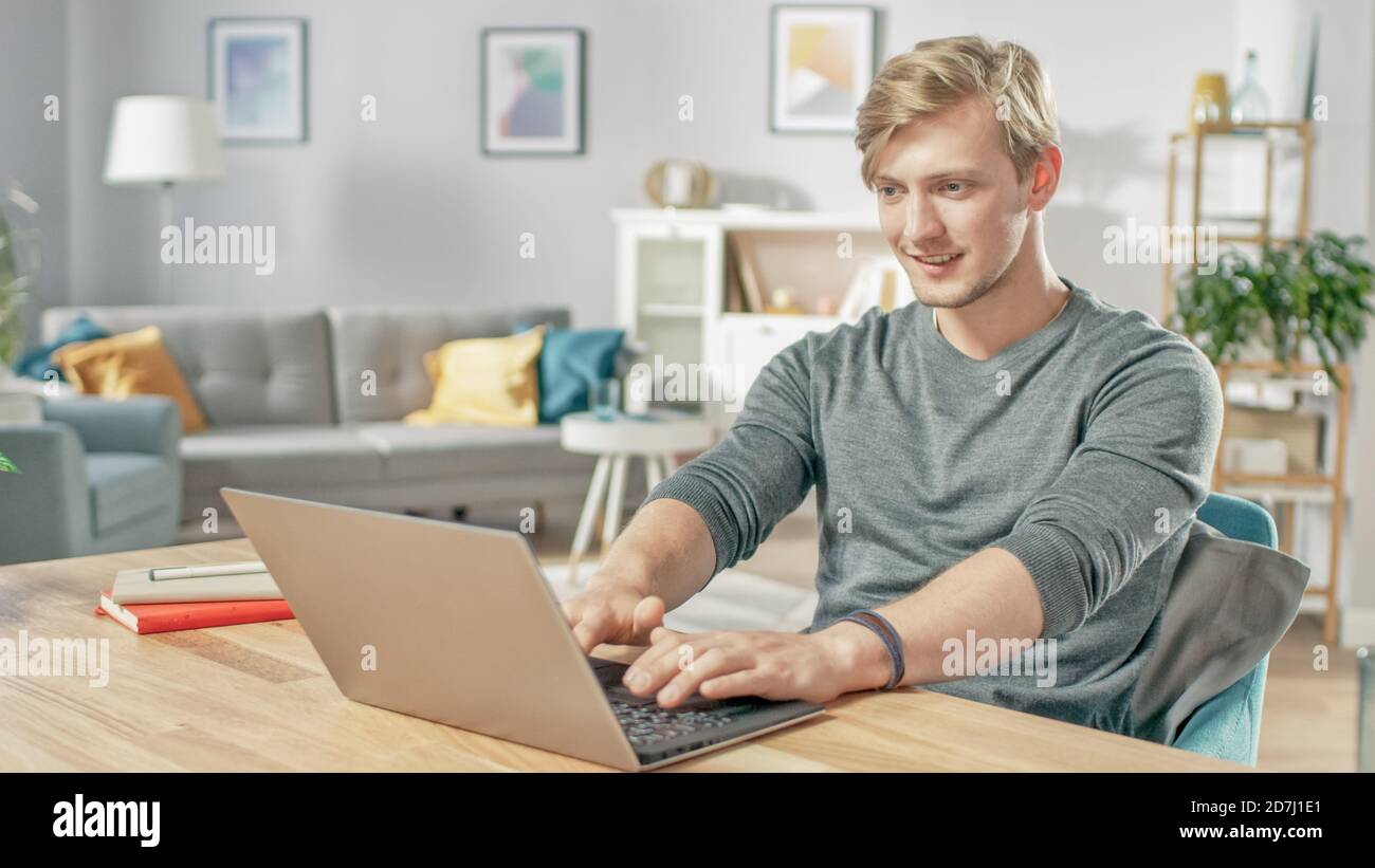 Bell'uomo giovane sorridente che usa il computer portatile mentre si siede alla scrivania del suo accogliente salotto. Foto Stock