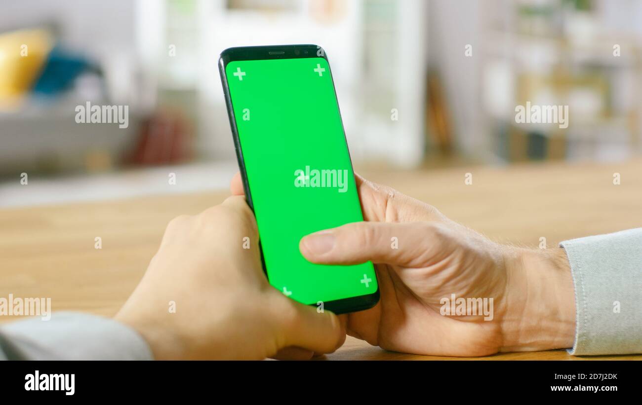 Primo piano di un uomo che tiene uno smartphone Green Mock-up schermo nelle sue mani. Utilizzo di un dispositivo wireless per navigare in Internet. In background accogliente Foto Stock