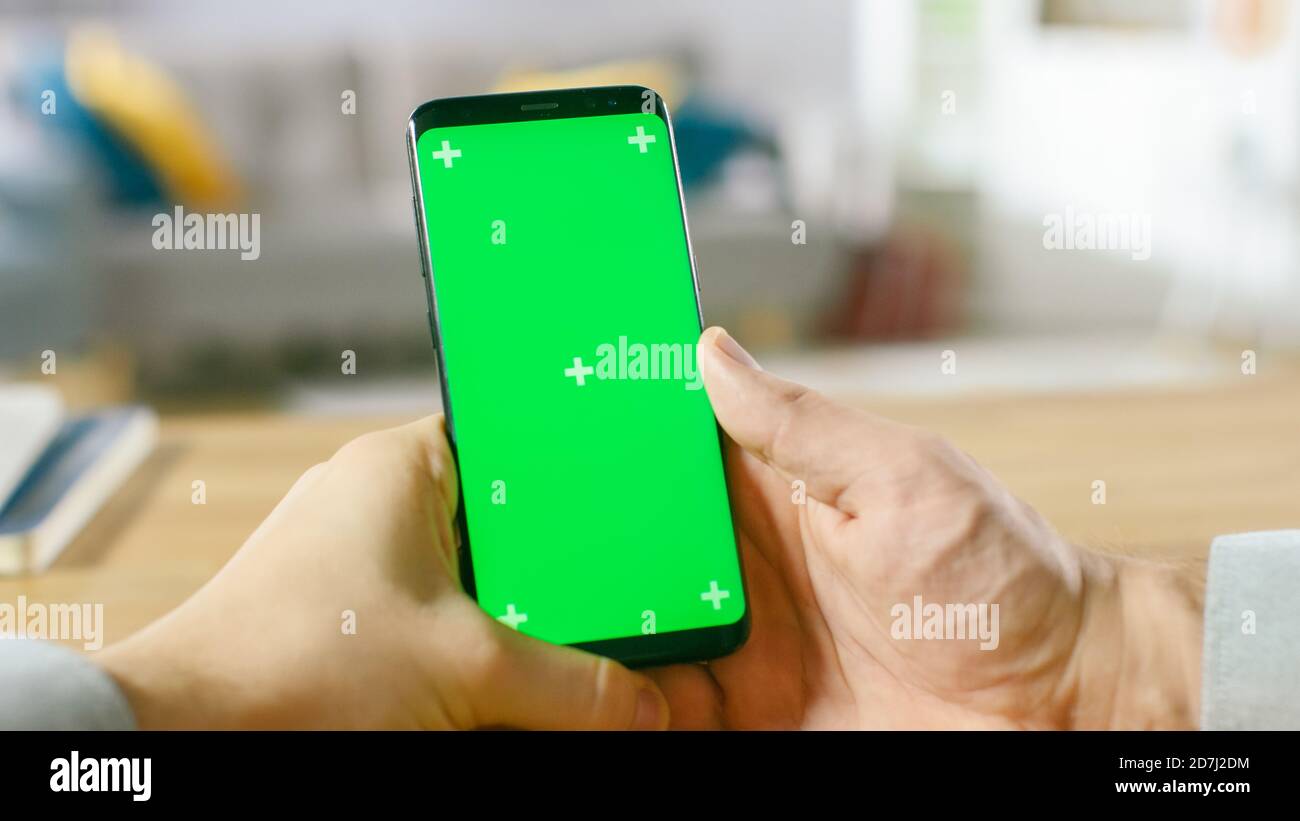 Primo piano di un uomo che tiene uno smartphone Green Mock-up schermo nelle sue mani. Utilizzo di un dispositivo wireless per navigare in Internet. In background accogliente Foto Stock
