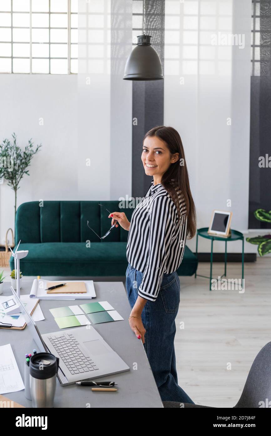 Sorridente bella giovane donna d'affari in piedi alla scrivania in un luogo di lavoro creativo Foto Stock