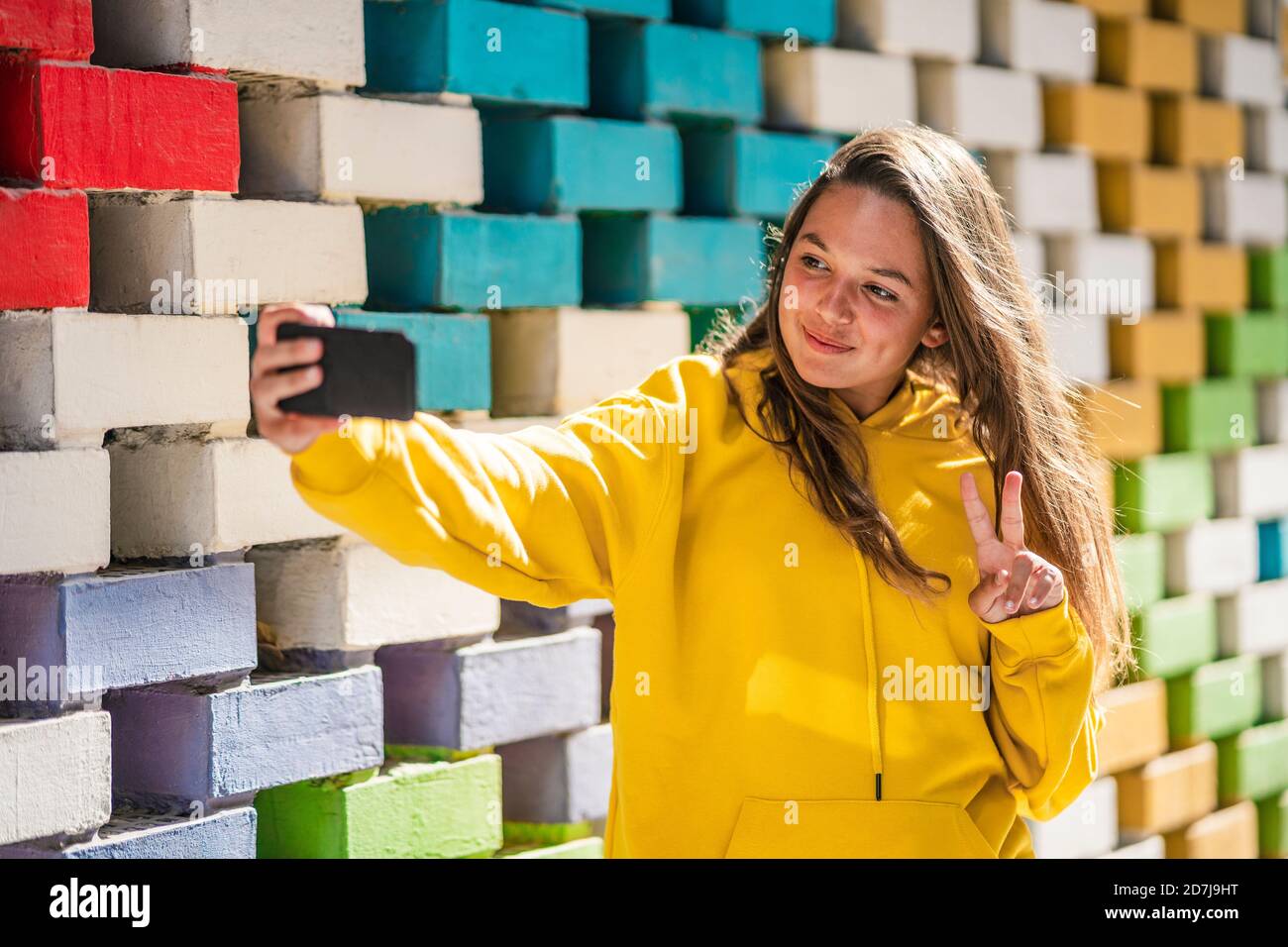 Giovane donna che prende selfie su astuto mentre si levano in piedi da colorato muro di mattoni Foto Stock