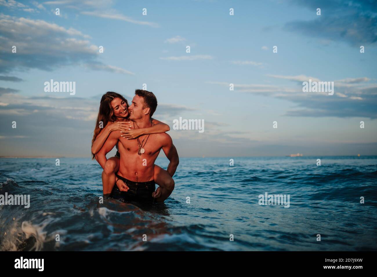 Uomo sorridente donna piggybacking mentre si trova in acqua in spiaggia Foto Stock