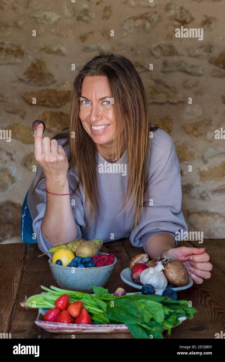 Felice dietista femminile con frutta e verdura varie sedute a. tavolo contro muro di pietra Foto Stock