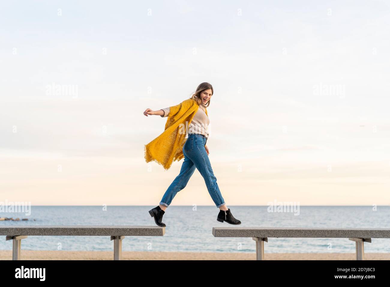 Giovane donna allegra che salta dalla panchina ad un'altra in spiaggia passeggiata contro il cielo durante il tramonto Foto Stock
