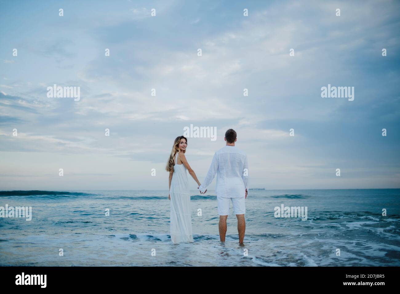 Donna che guarda dietro mentre tiene la mano dell'uomo in piedi dentro acqua in spiaggia Foto Stock