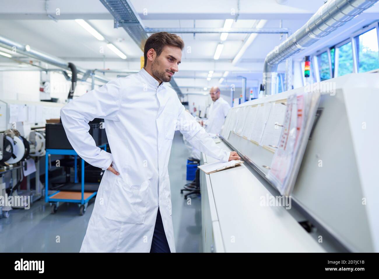 Giovane tecnico maschile in camice di laboratorio che guarda i rapporti su macchinari Foto Stock