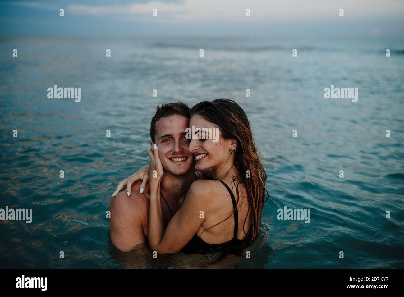 Una coppia sorridente che si abbraccia mentre si è in piedi in acqua in spiaggia durante tramonto Foto Stock